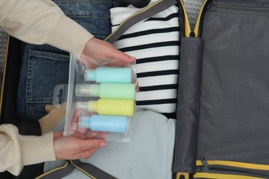 Photo of Woman with plastic bag of cosmetic travel kit packing suitcase, top view. Bath accessories