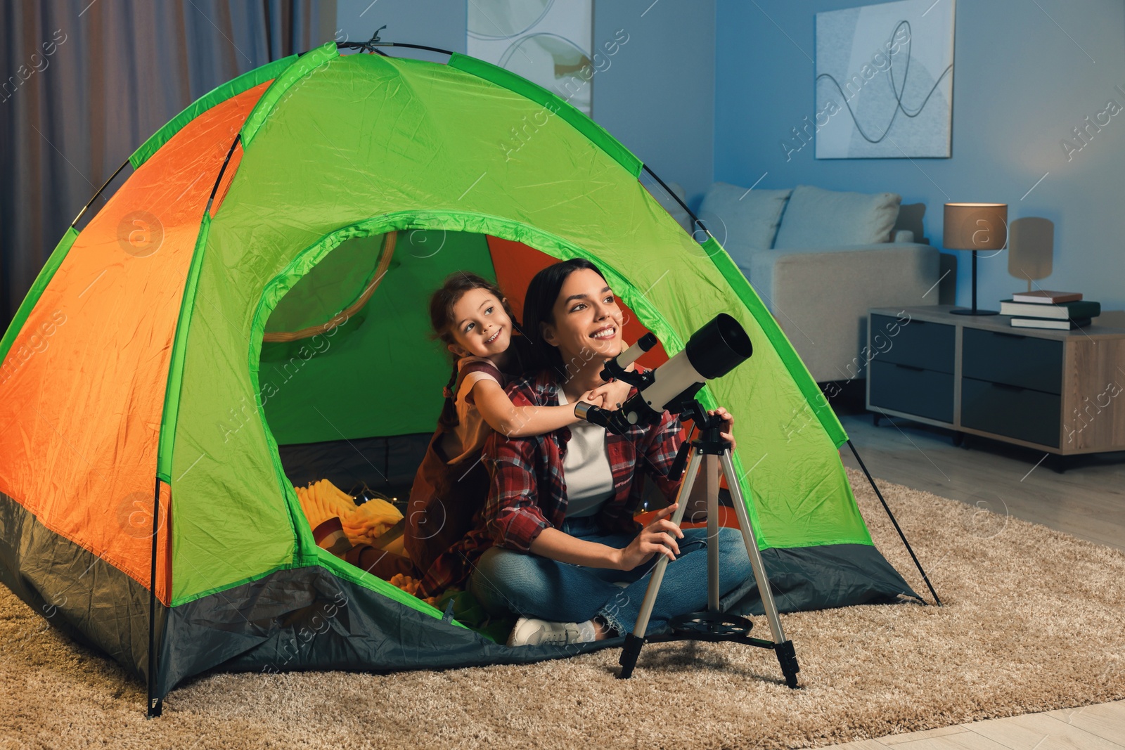 Photo of Happy mother and her daughter using telescope to look at stars while sitting in camping tent indoors