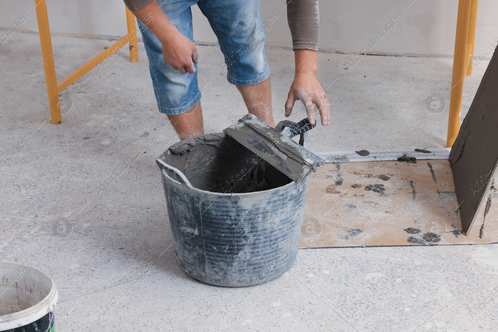 Photo of Worker taking spatula from bucket of adhesive mix near tile indoors, closeup