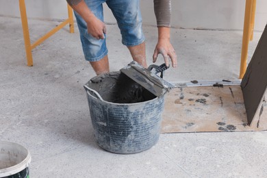 Photo of Worker taking spatula from bucket of adhesive mix near tile indoors, closeup