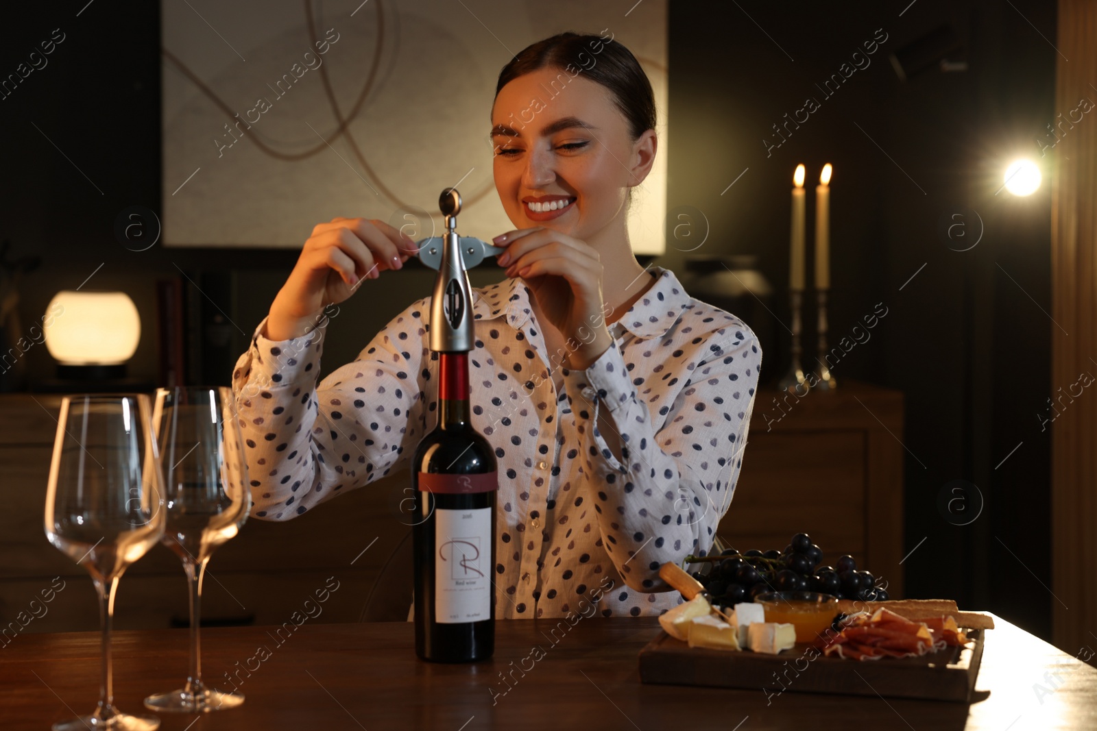 Photo of Romantic dinner. Happy woman opening wine bottle with corkscrew at table indoors