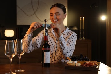Photo of Romantic dinner. Happy woman opening wine bottle with corkscrew at table indoors