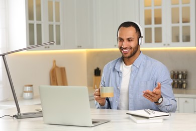 Photo of Young man with headphones having online video chat at desk in kitchen. Home office