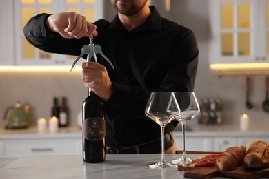 Romantic dinner. Man opening wine bottle with corkscrew at table in kitchen, closeup