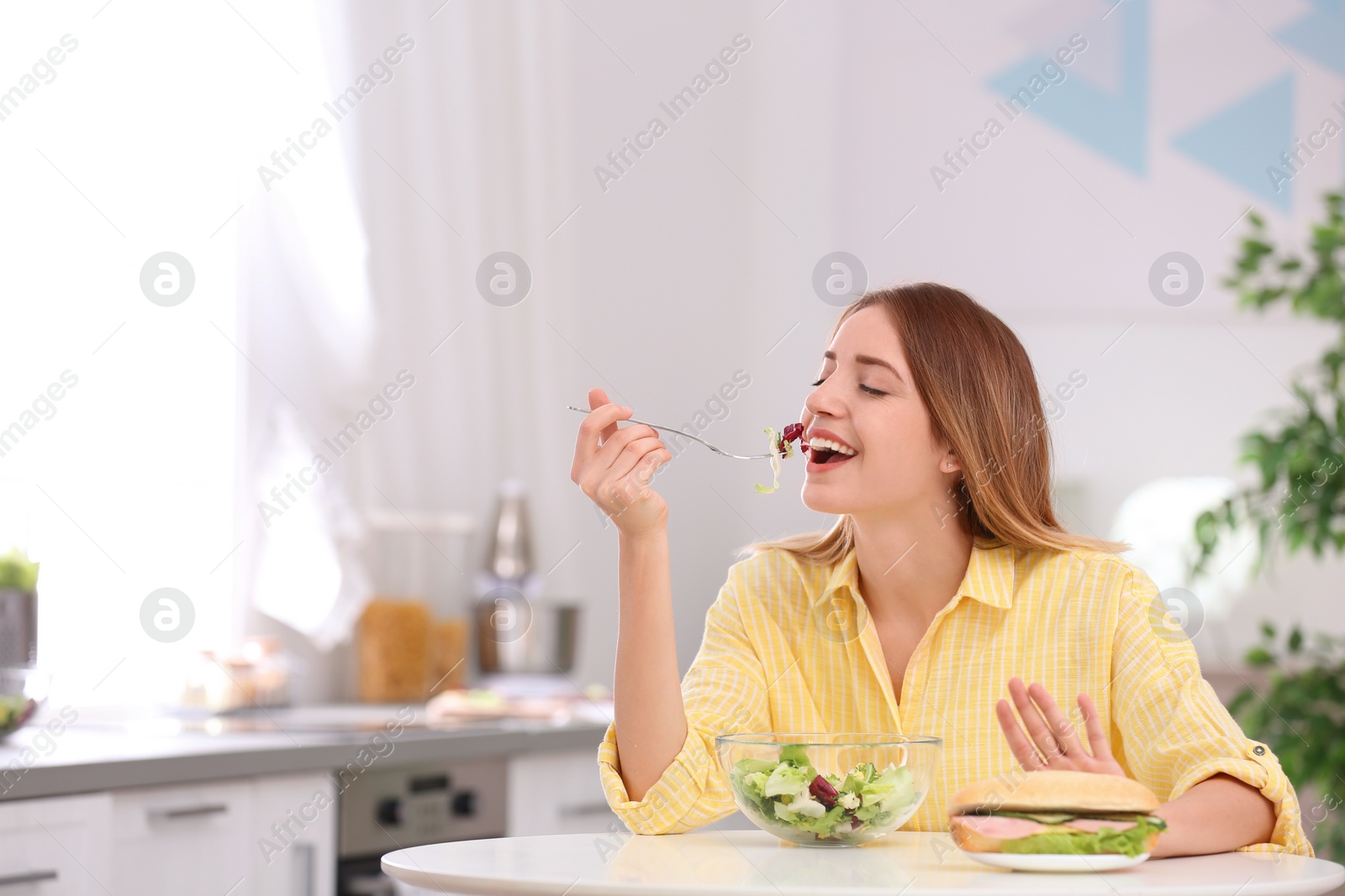 Photo of Happy young woman eating salad instead of sandwich in kitchen, space for text. Healthy diet
