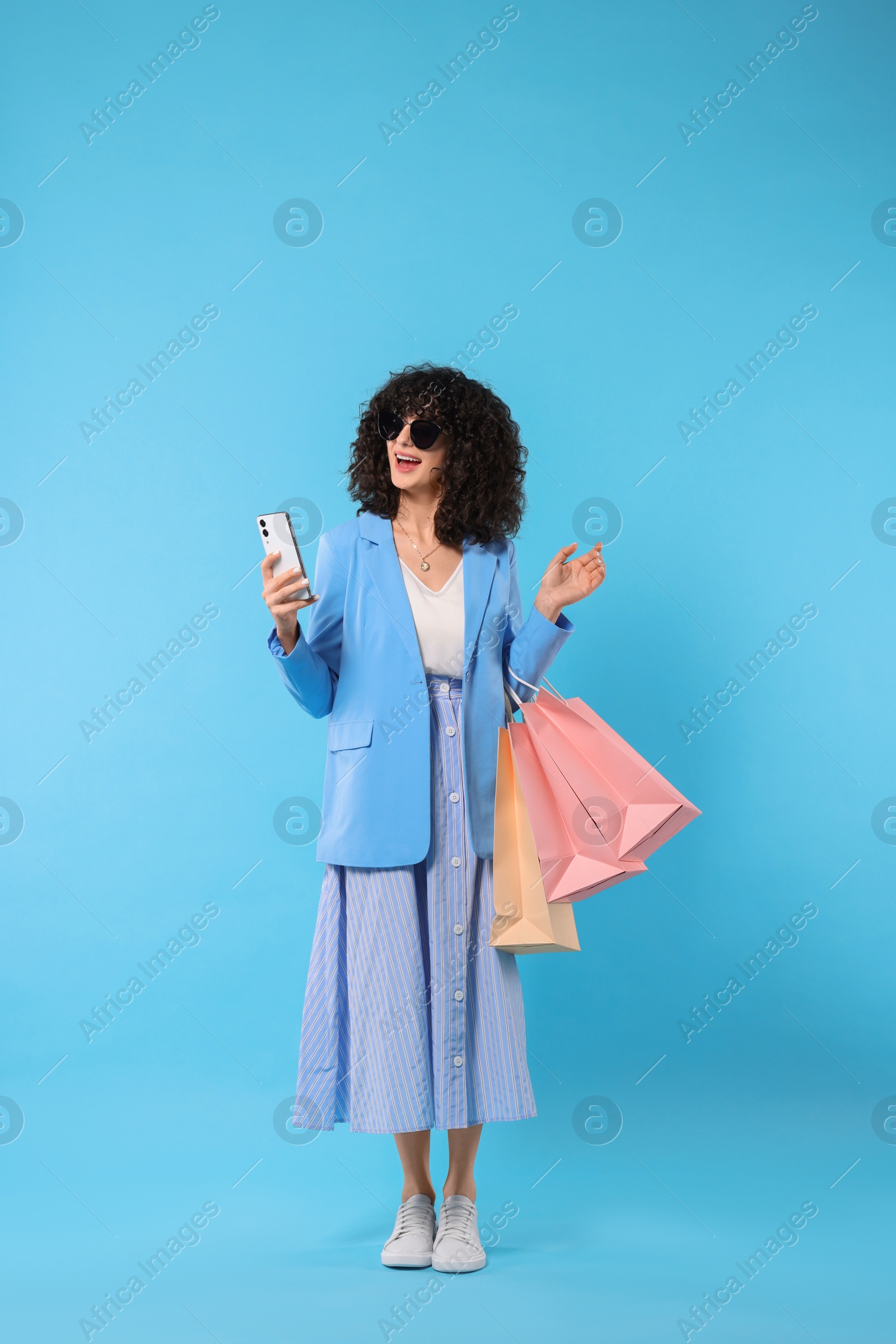 Photo of Happy young woman with shopping bags and modern smartphone on light blue background