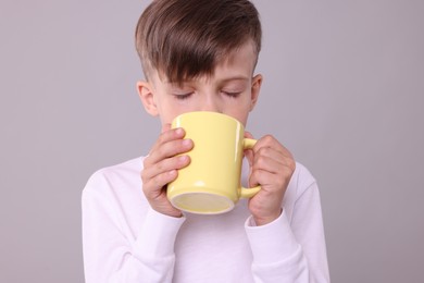 Cute boy drinking beverage from yellow ceramic mug on light grey background