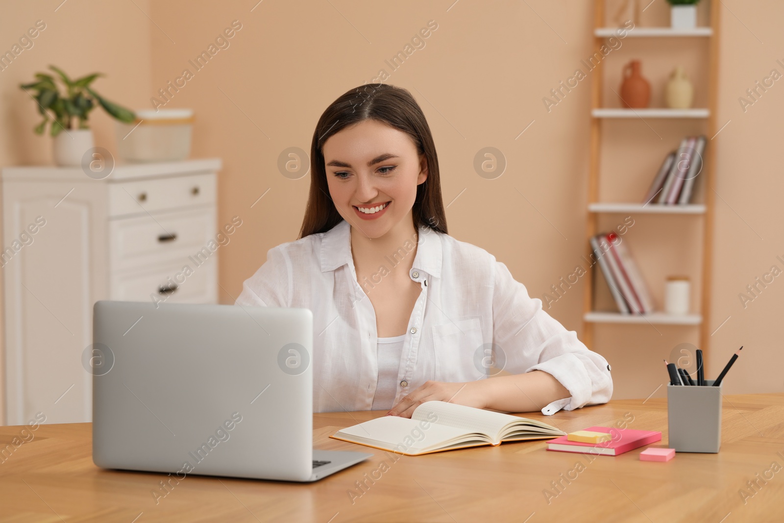 Photo of Happy young woman with notebook working on laptop at wooden table indoors