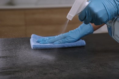 Photo of Woman in gloves cleaning grey stone table with rag and detergent indoors, closeup