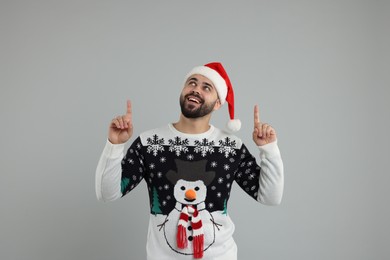 Photo of Happy young man in Christmas sweater and Santa hat pointing at something on grey background