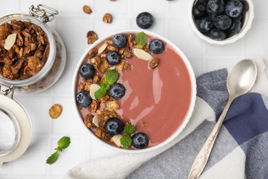 Photo of Bowl of delicious smoothie served with fresh blueberries and granola on white tiled table, flat lay