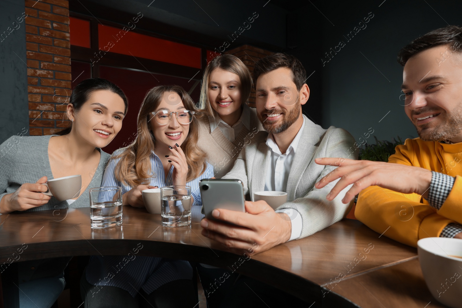 Photo of Handsome man showing something funny in smartphone to his friends in cafe