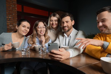 Photo of Handsome man showing something funny in smartphone to his friends in cafe