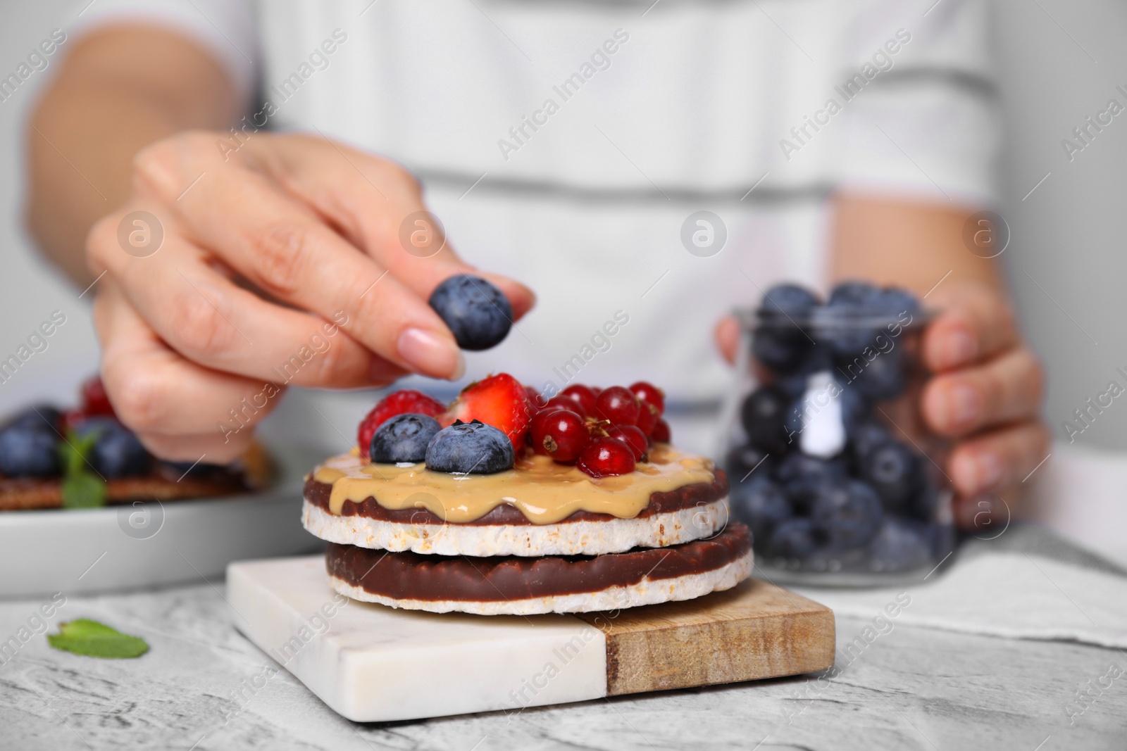 Photo of Woman decorating crunchy rice cakes with blueberry at white textured table, closeup