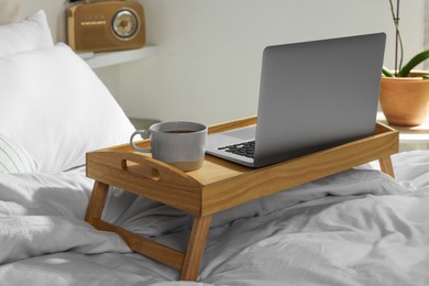 Wooden tray with modern laptop and cup of aromatic tea on bed indoors