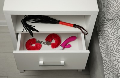 Photo of Bedside table with black leather whip, vibrator and fluffy handcuffs indoors. Sex toys