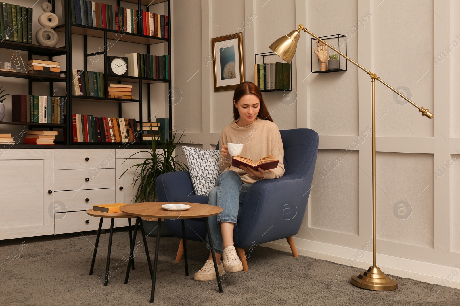 Photo of Young woman with cup of coffee reading book in armchair indoors. Home library