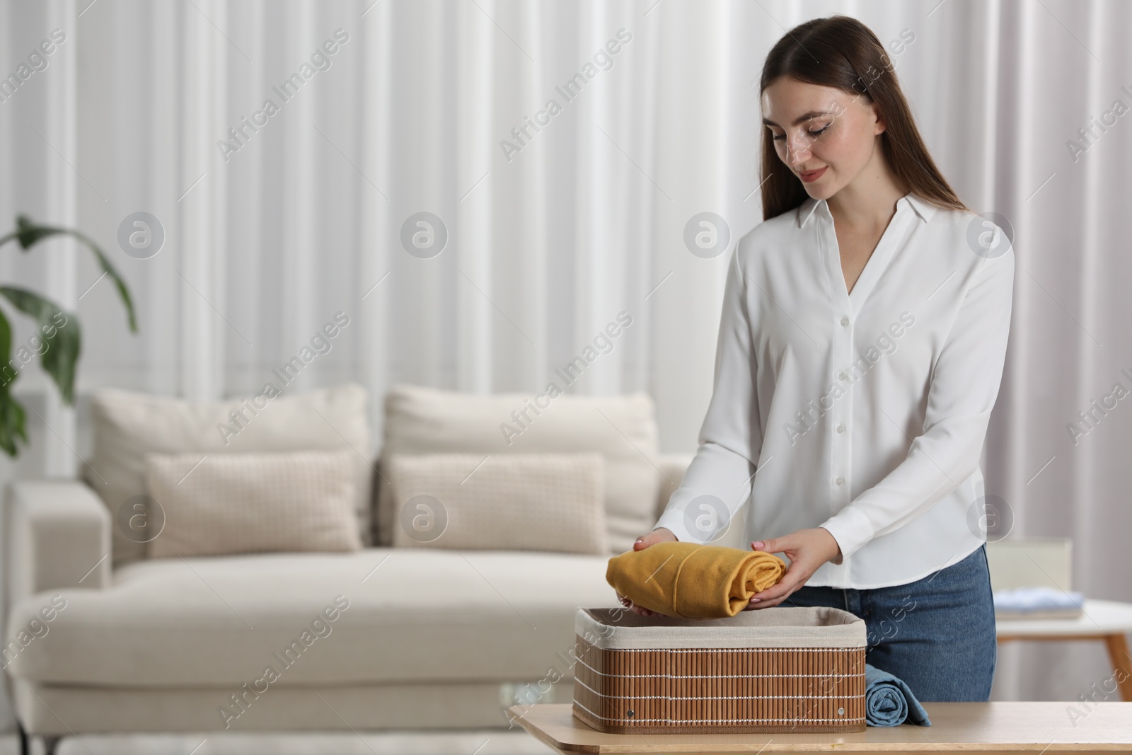 Photo of Young woman putting rolled shirt into basket at table in room, space for text. Organizing clothes