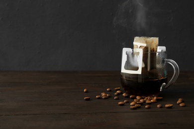 Photo of Glass cup with drip coffee bag and beans on black wooden table. Space for text
