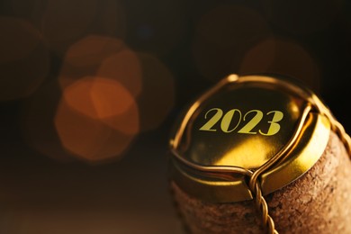 Cork of sparkling wine and muselet cap with engraving 2023 against blurred festive lights, closeup. Bokeh effect