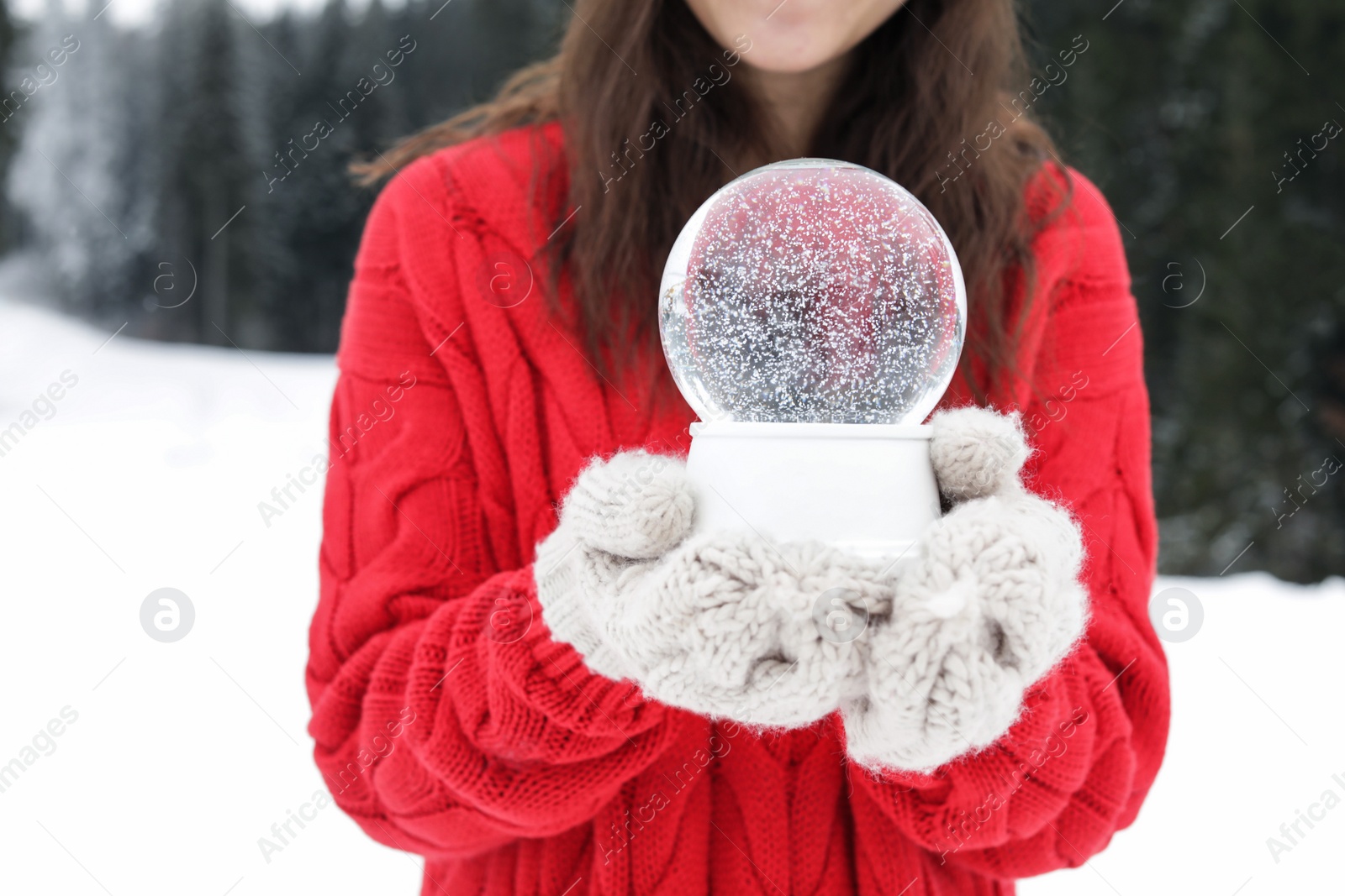 Photo of Woman with knitted mittens holding snow globe outdoors, closeup. Winter vacation