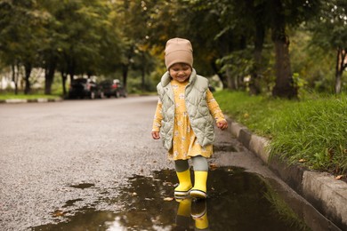 Photo of Cute little girl splashing water with her boots in puddle outdoors, space for text