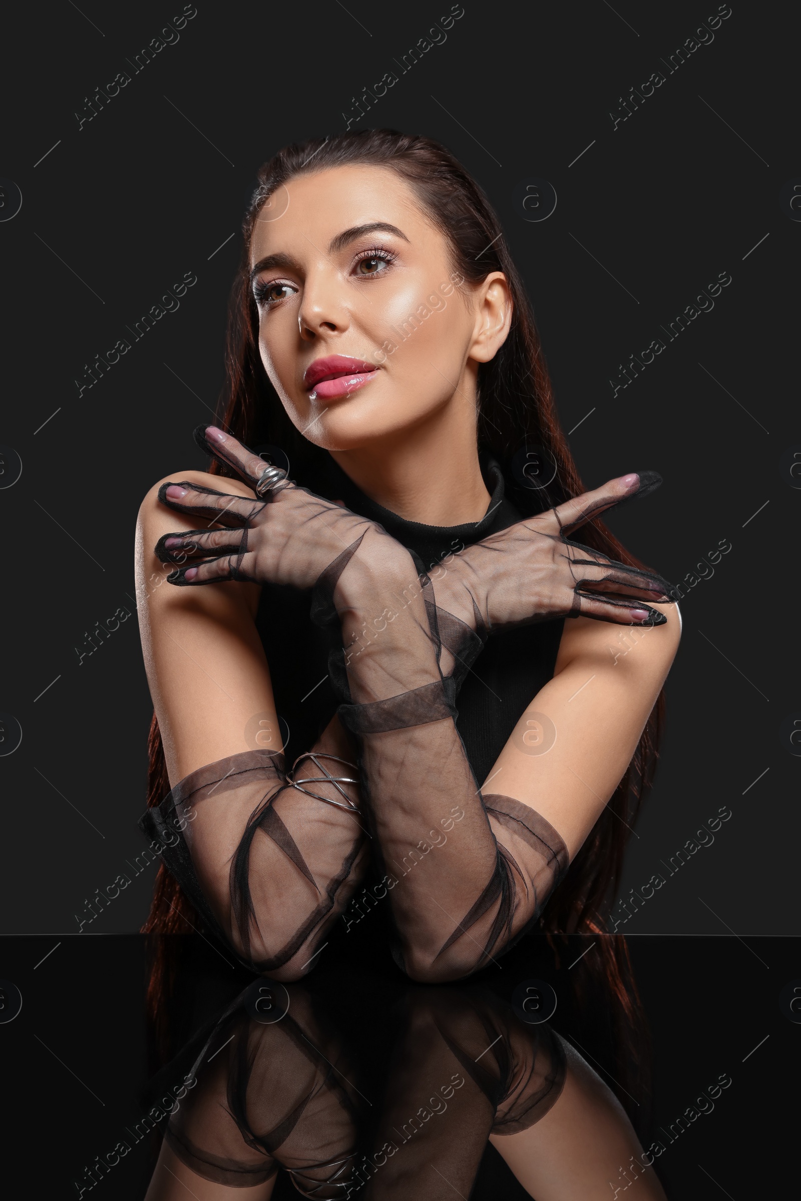 Photo of Beautiful young woman in evening gloves at black glass table against dark background