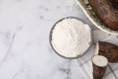 Bowl with cassava flour and roots on white marble table, flat lay. Space for text