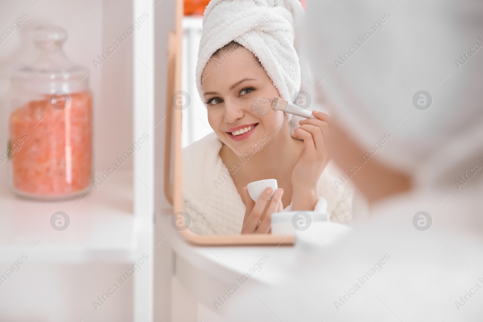 Photo of Woman applying scrub onto face in front of mirror at home