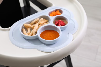 Photo of High chair with food in baby tableware on tray indoors, closeup