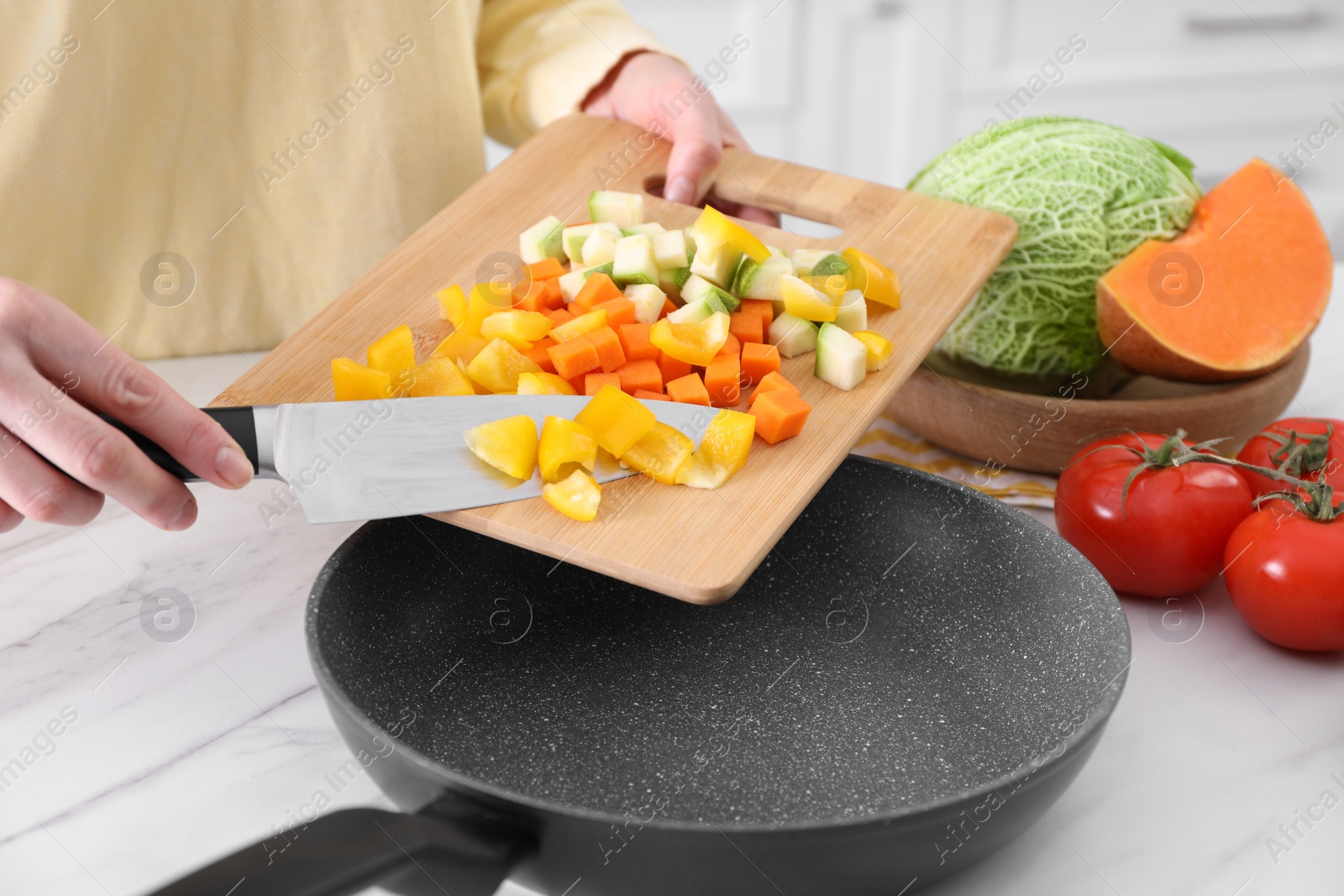 Photo of Woman pouring mix of cut vegetables into frying pan at table in kitchen, closeup