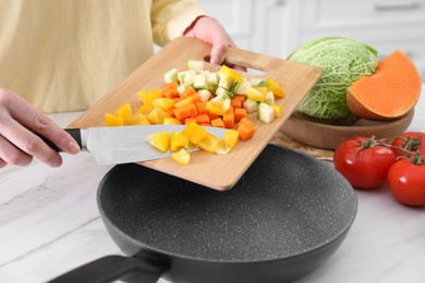 Woman pouring mix of cut vegetables into frying pan at table in kitchen, closeup