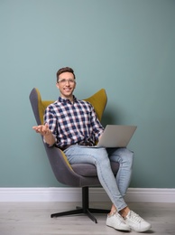 Young blogger with laptop sitting in armchair against color wall