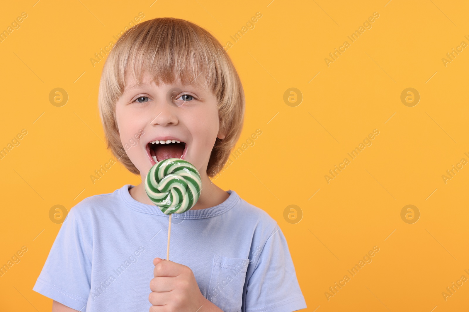 Photo of Happy little boy licking bright lollipop swirl on orange background, space for text