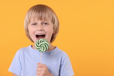 Photo of Happy little boy licking bright lollipop swirl on orange background, space for text