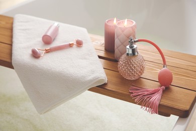 Photo of Wooden bath tray with face roller, cosmetic products and candles on tub indoors