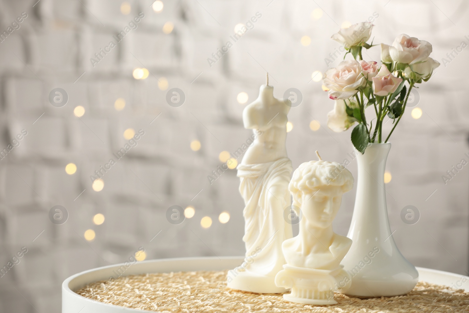 Photo of Beautiful David bust and Venus De Milo candles near flowers on table against blurred lights. Space for text