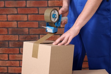 Photo of Worker taping box with adhesive tape dispenser near brick wall, closeup
