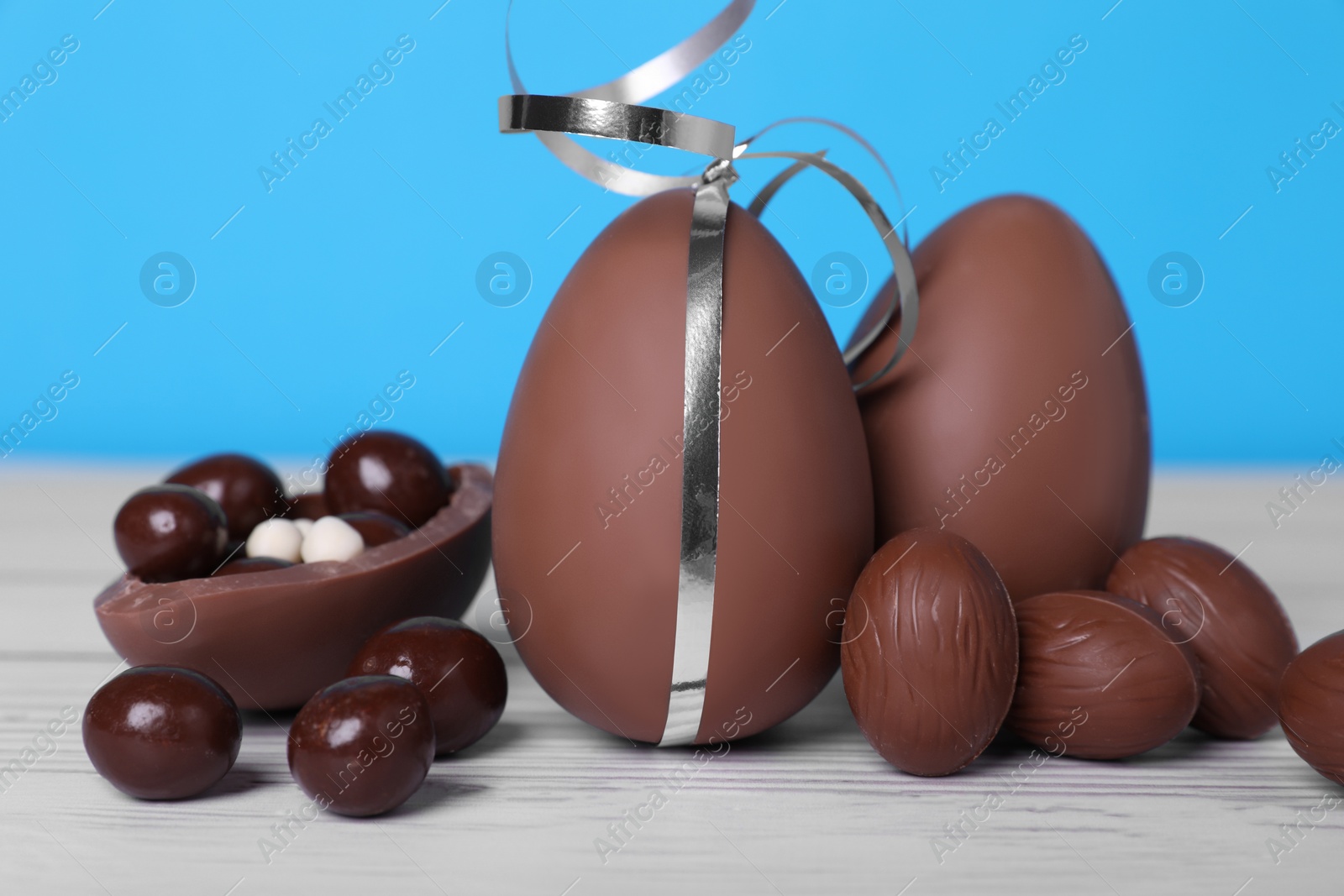 Photo of Delicious chocolate eggs and candies on white wooden table against light blue background, closeup