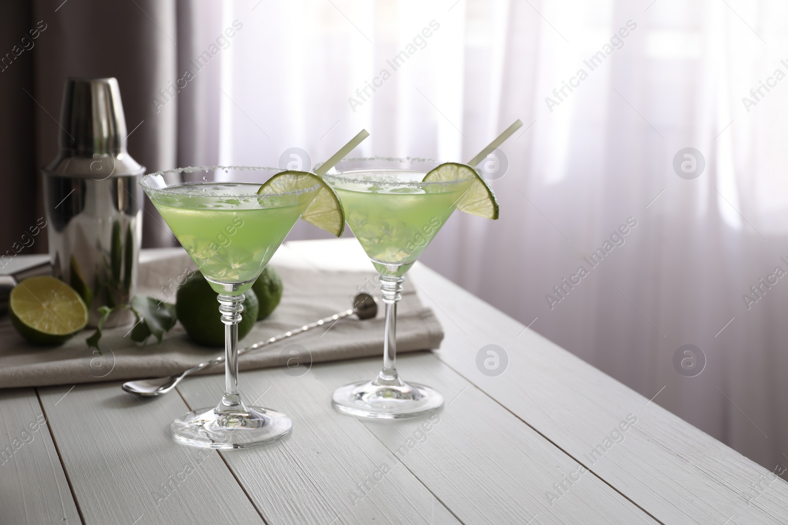 Photo of Delicious Margarita cocktail in glasses, limes, bar spoon and shaker on white wooden table, space for text