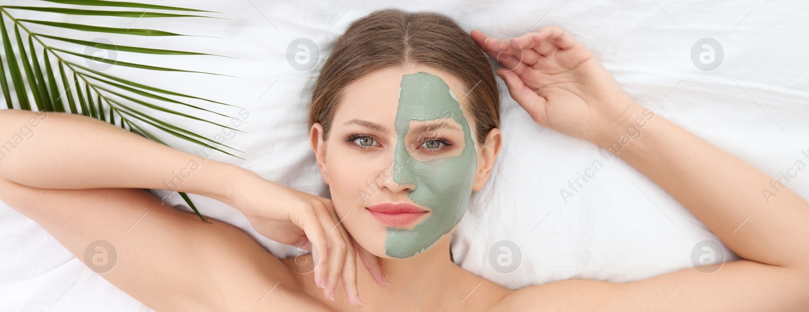Image of Beautiful woman with clay facial mask and tropical leaf on white fabric, above view. Banner design