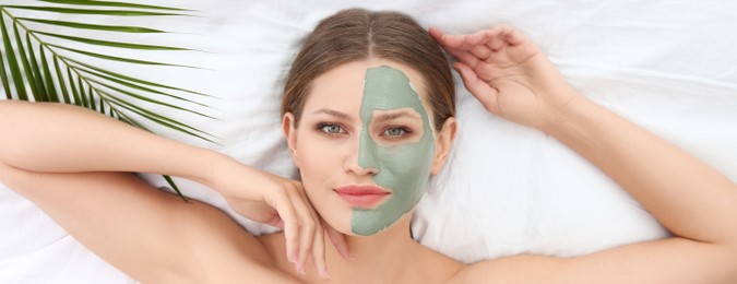 Beautiful woman with clay facial mask and tropical leaf on white fabric, above view. Banner design