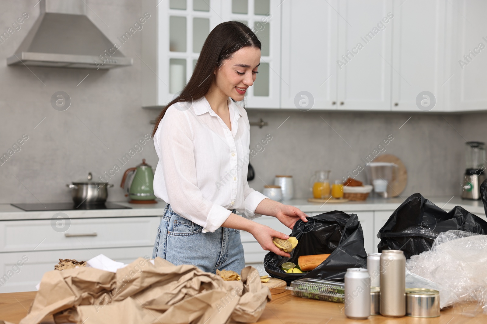 Photo of Garbage sorting. Woman putting food waste into plastic bag at table in kitchen