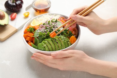 Photo of Woman eating delicious vegan bowl with avocados, carrots and tomatoes at light table, closeup