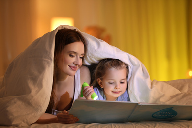 Photo of Mother and daughter with flashlight reading book under blanket at home