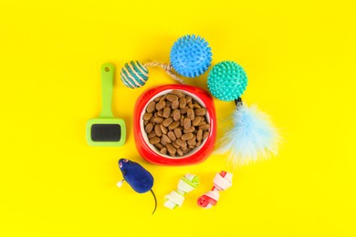Photo of Bowl of dry food, brush and toys on yellow background, flat lay. Pet shop goods