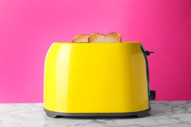Yellow toaster with roasted bread on white marble table against pink background