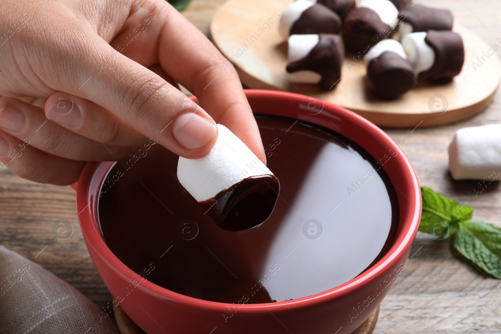 Photo of Woman dipping marshmallow into melted chocolate at wooden table, closeup