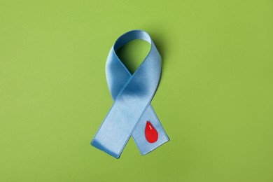 Photo of Light blue ribbon with paper blood drop on green background, top view. Diabetes awareness