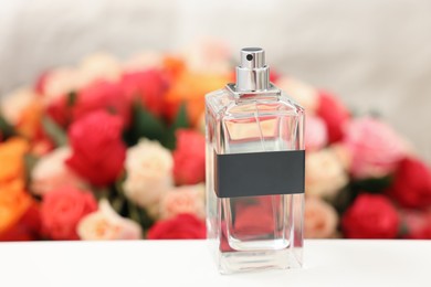 Bottle of perfume on white table against bouquet of beautiful roses, closeup. Space for text